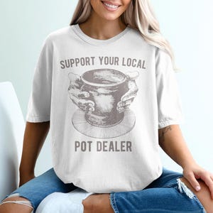 May include: White t-shirt with the text "SUPPORT YOUR LOCAL POT DEALER" in gray. The shirt features a gray illustration of a pot being held by hands. The shirt is a casual, comfortable style.
