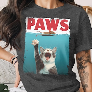 May include: Dark gray t-shirt featuring a playful cat illustration. The image shows a cat reaching for a mouse swimming above the water line. The word "PAWS" is printed in red above the cat. The shirt is made of a soft material.
