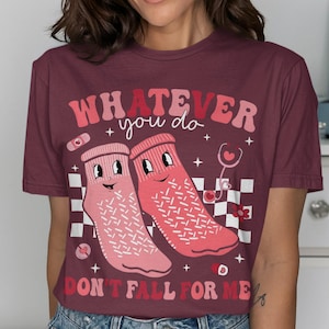 May include: Burgundy t-shirt with the text "WHATEVER you do DON'T FALL FOR ME". The graphic features two smiling socks, a stethoscope, and a bandage. The design is in shades of pink and red.