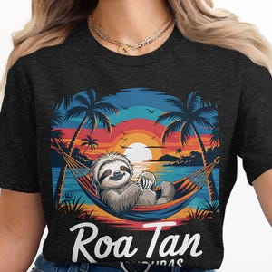 May include: Dark gray t-shirt featuring a sloth relaxing in a hammock on a tropical beach. The design includes palm trees, a sunset, and the text "Roa Tan". The sloth is smiling, and the overall theme is relaxation and vacation.