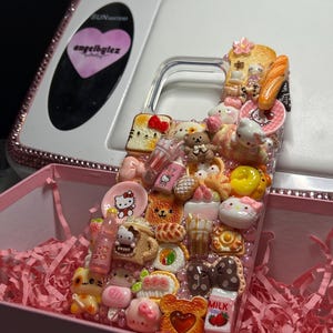 May include: A clear phone case decorated with numerous miniature food and character charms, including Hello Kitty figures, toast, pastries, milk cartons, and small pink pearls. The charms are arranged in a dense, layered collage across the surface.