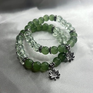 May include: Two green beaded bracelets with silver marijuana leaf charms. The bracelets are made of green and clear beads.
