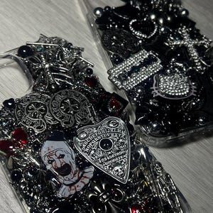 May include: Two clear phone cases heavily embellished with gothic and horror-themed charms. One case features a Ouija board, a clown, and a Scream mask. The other case has a Hello Kitty charm and various black and silver accents.