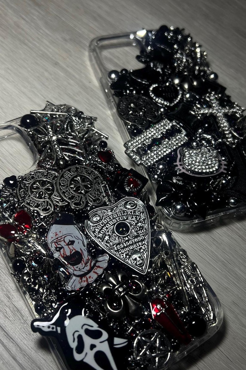 May include: Two clear phone cases heavily embellished with gothic and horror-themed charms. One case features a Ouija board, a clown, and a Scream mask. The other case has a Hello Kitty charm and various black and silver accents.