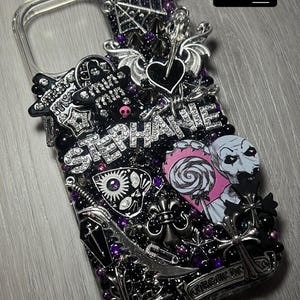 May include: A clear phone case heavily embellished with black and silver gothic-themed charms, including a spiderweb, hearts, and crosses. The name "STEPHANIE" is spelled out in silver rhinestones. The case also features a pink and white lollipop and a zombie-like illustration.