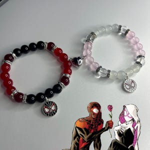 May include: Two beaded bracelets. One bracelet features red and black beads with a Spider-Man charm. The other bracelet has pink and clear beads with a Spider-Man charm. Both bracelets have silver accents and a heart-shaped magnetic clasp.
