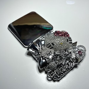 Op de afbeelding: Een zilveren compacte spiegel met een open deksel. De compact is versierd met een sprankelend, met juwelen bezet ontwerp met een strik, een kattengezicht en andere versieringen. De kattenkop is versierd met roze edelstenen.