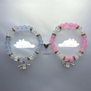 Puede incluir: Dos pulseras a juego con un dije de corazón que las conecta. Una pulsera tiene cuentas de color azul claro y la otra tiene cuentas de color rosa. Ambas pulseras tienen cuentas de cristal transparente y un dije de un perro de dibujos animados.
