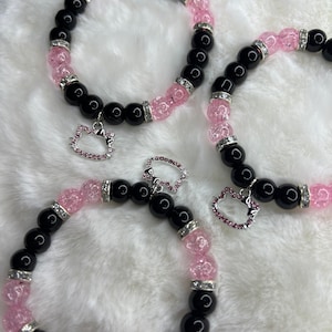 May include: Three black and pink beaded bracelets with a silver charm in the shape of a cat head. Each charm has pink rhinestones.