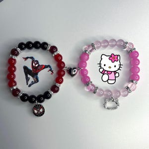 May include: Two beaded bracelets connected by a silver heart charm. The bracelet on the left has red and black beads with a Spiderman charm. The bracelet on the right has pink beads with a Hello Kitty charm.