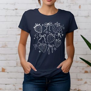 K&ouml;nnte beinhalten: Marineblaues T-Shirt mit wei&szlig;em Umrissdesign von Erdbeeren, Schleifen und Sternen. Das kurz&auml;rmelige Shirt ist aus einem weichen Material gefertigt. Das Design ist mittig auf der Vorderseite des Shirts angeordnet.