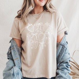 K&ouml;nnte beinhalten: Beiges T-Shirt mit wei&szlig;em Erdbeer- und Schleifendesign. Die Grafik zeigt mehrere Erdbeeren und Schleifen sowie kleine sternf&ouml;rmige Akzente. Das Shirt hat kurze &Auml;rmel und einen Rundhalsausschnitt.