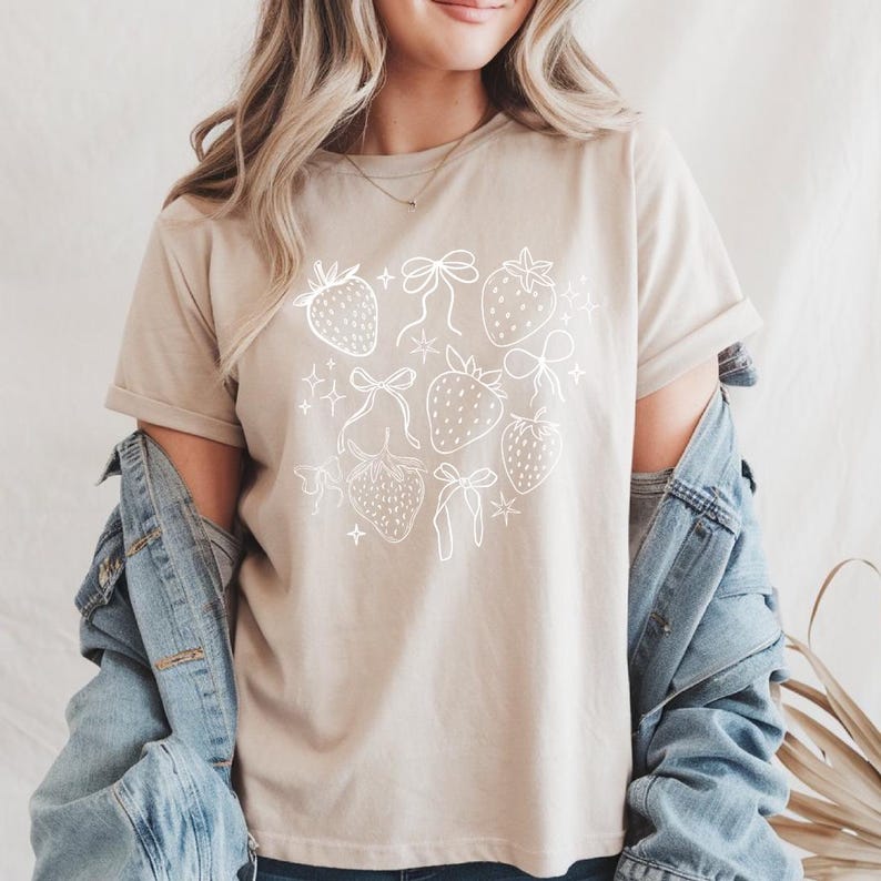 K&ouml;nnte beinhalten: Beiges T-Shirt mit wei&szlig;em Erdbeer- und Schleifendesign. Die Grafik zeigt mehrere Erdbeeren und Schleifen sowie kleine sternf&ouml;rmige Akzente. Das Shirt hat kurze &Auml;rmel und einen Rundhalsausschnitt.