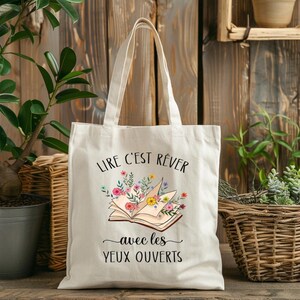 Peut inclure: Sac fourre-tout crème avec l'inscription française "LIRE C'EST RÊVER avec les YEUX OUVERTS" et une illustration d'un livre fleuri. Le sac a deux anses et est fait d'un tissu résistant.