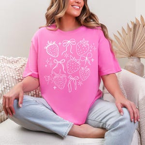 K&ouml;nnte beinhalten: Ein leuchtend pinkfarbenes T-Shirt mit einer wei&szlig;en Grafik von Erdbeeren, Schleifen und Sternen. Das Design ist mittig auf der Vorderseite platziert. Das T-Shirt hat einen Rundhalsausschnitt und kurze &Auml;rmel.