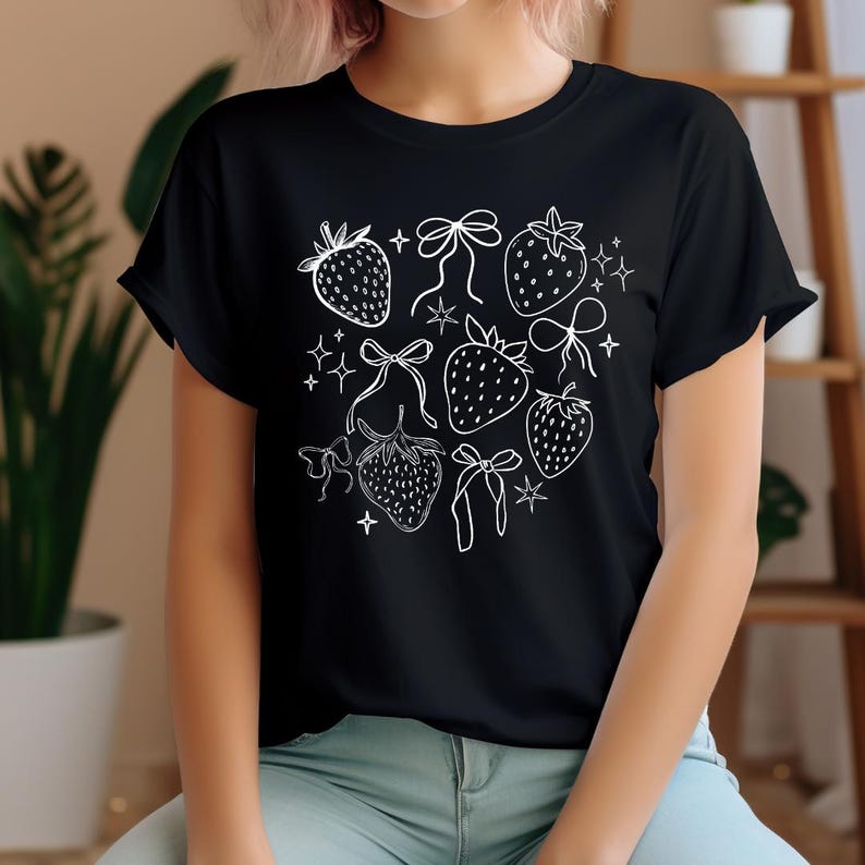 K&ouml;nnte beinhalten: Schwarzes T-Shirt mit einer wei&szlig;en Strichzeichnung von Erdbeeren, Schleifen und Sternen. Das Design befindet sich mittig auf dem Shirt. Das T-Shirt hat einen Rundhalsausschnitt und kurze &Auml;rmel. Das Design ist schlicht und elegant.
