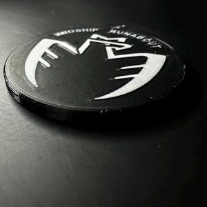 May include: A black, circular emblem with white lettering and a stylized wing design. The text "MIDSHIP RUNABOUT" is visible around the edge. The emblem is slightly raised, creating a 3D effect.