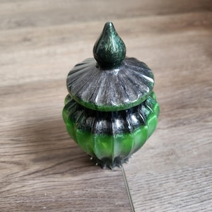 May include: A decorative jar with a lid, featuring a green and silver color scheme. The jar has a rounded, ribbed body and a matching lid with a pointed finial. The color transitions from green to silver on the lid.