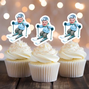 Puede incluir: Tres cupcakes blancos con glaseado azul y blanco cubiertos con una imagen impresa de un niño con un traje de esquí azul y gafas de esquí bajando una montaña nevada.