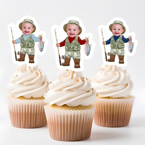 May include: Three cupcakes decorated with images of a baby dressed as a fisherman holding a fishing rod and a fish. The cupcakes are frosted with white frosting and have gold sprinkles.
