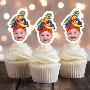 Op de afbeelding: Drie cupcakes met witte frosting en eetbare fruit- en babygezicht-toppers. De toppers tonen een babygezicht met een fruitmuts.