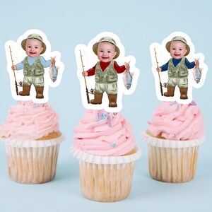 May include: Three cupcake toppers featuring a cartoon baby dressed as a fisherman holding a fishing rod and a fish. The baby is wearing a green vest, brown pants, and brown boots. The toppers are on pink cupcakes with white frosting and sprinkles.