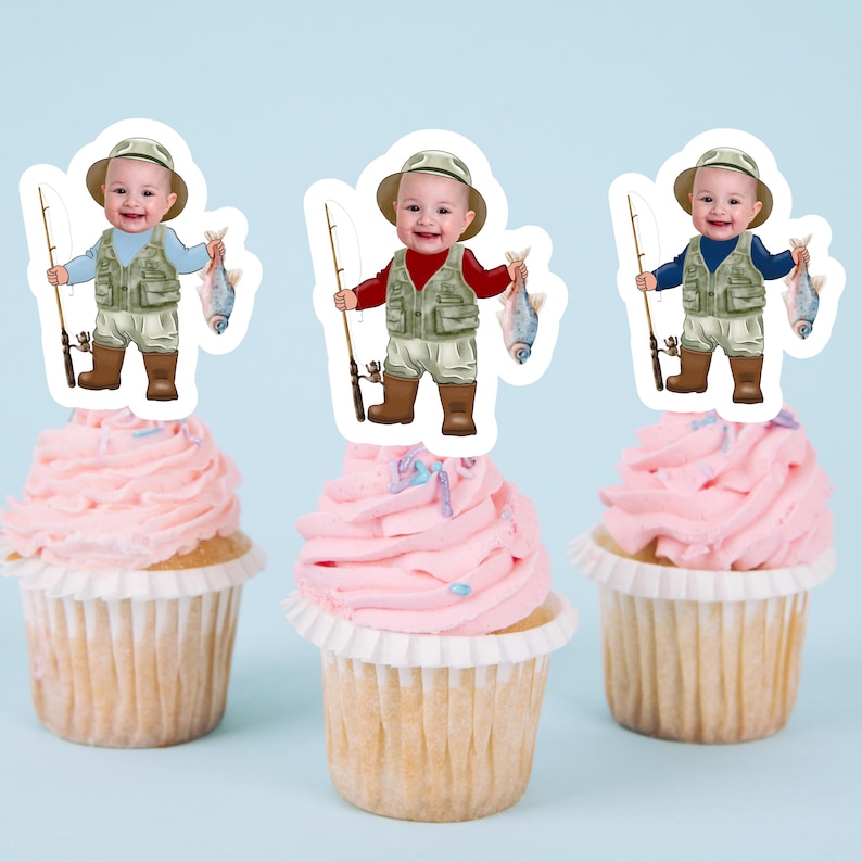 May include: Three cupcake toppers featuring a cartoon baby dressed as a fisherman holding a fishing rod and a fish. The baby is wearing a green vest, brown pants, and brown boots. The toppers are on pink cupcakes with white frosting and sprinkles.