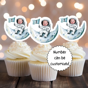 May include: Three white cupcakes with white frosting and pearl sprinkles topped with astronaut themed cupcake toppers. The toppers feature a crescent moon with a cartoon astronaut holding a flag with the number "1" on it. The text "Number can be customized" is visible on a white circle.