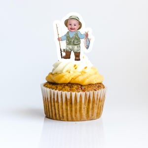 May include: A cupcake topped with a white frosting and a  topper of a cartoon baby boy wearing a fishing vest and holding a fishing rod and a fish.