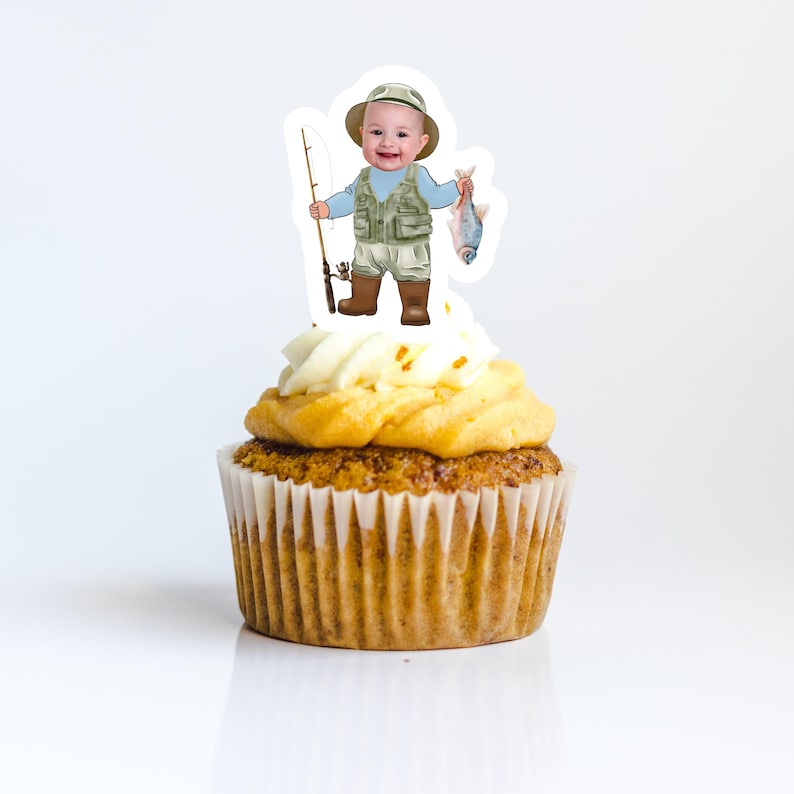 May include: A cupcake topped with a white frosting and a  topper of a cartoon baby boy wearing a fishing vest and holding a fishing rod and a fish.