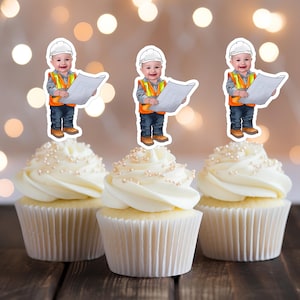 May include: Three white cupcakes with white frosting and pearl sprinkles topped with construction worker themed cupcake toppers. Each topper features a cartoon image of a young boy wearing a white hard hat, orange vest, blue jeans, and holding a blueprint.