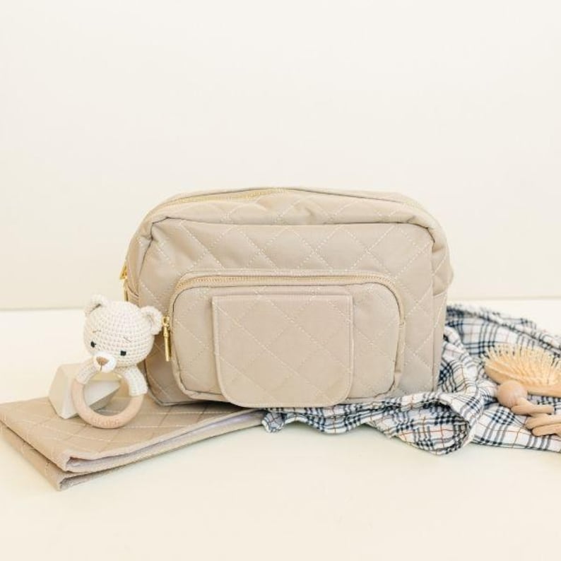 May include: A beige quilted diaper bag with a gold zipper and a large front pocket. A small crocheted teddy bear with a wooden ring is in the foreground.