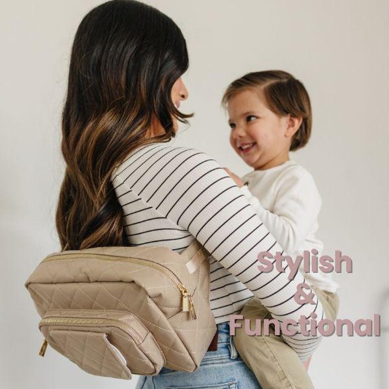 May include: A woman wearing a striped shirt is holding a toddler. She is wearing a beige quilted diaper bag with gold accents. The bag is worn as a hip pack. The text "Stylish & Functional" is superimposed on the image.