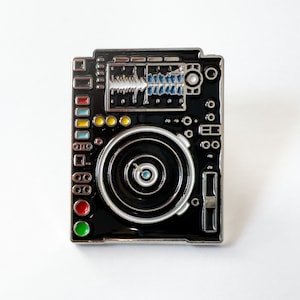 Puede incluir: Pin de esmalte negro y plateado de una mesa de mezclas de DJ con un tocadiscos en blanco y negro. El pin tiene detalles en rojo, amarillo, verde y azul.