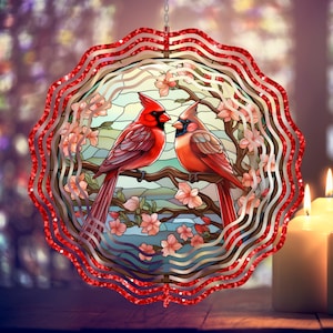 May include: A red and silver metal wind spinner with a stained glass design of two cardinals perched on a branch with pink flowers. The wind spinner has a red glitter finish.