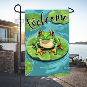 Puede incluir: Una bandera de jardín con una rana de dibujos animados con ojos rojos sentada en una hoja de nenúfar. La palabra "Welcome" está escrita encima. El fondo es un estanque azul con hojas de nenúfar.