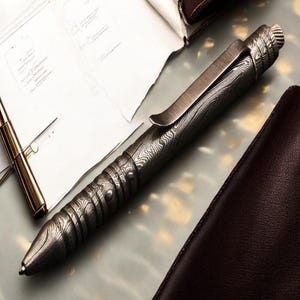 May include: A detailed close-up of a patterned metal pen with a clip, lying on a light-colored surface. The pen has a textured design and a silver finish. A gold pen and a brown leather wallet are also visible.