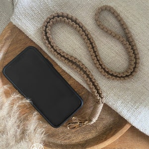 Könnte beinhalten: Ein beigefarbenes Makramee-Handyband mit einem goldfarbenen Verschluss wird neben einem schwarzen Smartphone in einer Hülle gezeigt. Das Band liegt auf einem Holztablett mit beigem Stoffhintergrund. Das Logo des Shops ist sichtbar.