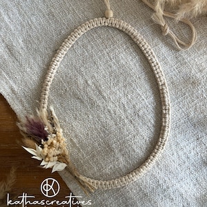 Könnte beinhalten: Ein beiger Makramee-Wandbehang mit einem Trockenblumenarrangement. Das kreisförmige Design zeigt einen geflochtenen Seilrahmen und ein kleines Bündel Trockenblumen in Lila-, Beige- und Weißtönen. Der Behang wird auf einem strukturierten, neutralfarbenen Stoff präsentiert.