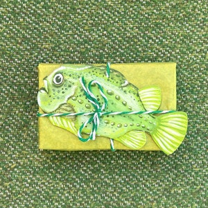 Könnte beinhalten: Eine Illustration eines grünen und weißen Fisches, verpackt in gelbes Papier mit grünem und weißem Garn.