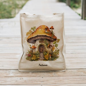 May include: A beige tote bag with a mushroom house illustration. The mushroom is orange and white with brown accents. The house has a door and windows. The bag has the word "Autumn" printed on the bottom.