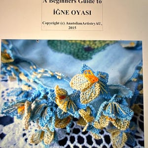 Op de afbeelding: Een close-up van een antieke zijden viooltjes ova in blauw en geel, een type Turks naaldkant. De kant is delicaat en ingewikkeld, met kleine bloemen en bladeren. De tekst "A Beginners Guide to İĞNE OYASI" en "Copyright (c) AnatolianArtistryAU, 2015" is zichtbaar bovenaan de afbeelding.