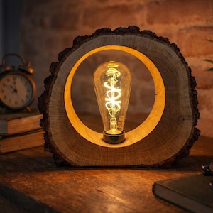Può includere: Una lampada da tavolo circolare realizzata con una spessa fetta di legno naturale con corteccia visibile sul bordo esterno. Il centro presenta una lampadina Edison in vetro trasparente con un filamento luminoso su una base scura.