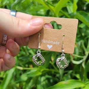 May include: Silver-colored earrings with a fang design, hanging from a brown tag that reads "Handmade" with a heart symbol. The earrings are held against a backdrop of green foliage.