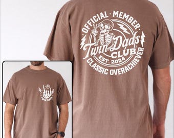Könnte beinhalten: Braunes T-Shirt mit weißem Aufdruck auf der Rückseite mit der Aufschrift "OFFICIAL MEMBER Twin Dads CLUB CLASSIC OVERACHIEVER EST. 2024" und einer Skelett-Illustration. Auf der Vorderseite befindet sich ein kleinerer weißer Aufdruck mit einer Handgeste und den Worten "Twin Dads".