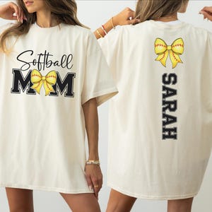 A woman wearing a white t-shirt with the text "Softball Mom" and a yellow bow on the back.