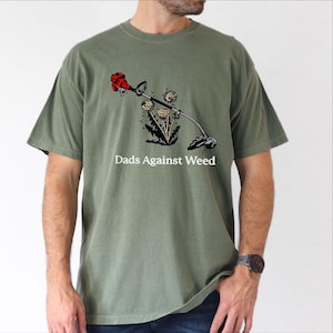 May include: Olive green t-shirt with a graphic of a weed whacker and dandelions. The text "Dads Against Weed" is printed below the graphic. The shirt is a short-sleeved crew neck.