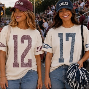 May include: Two beige short-sleeve t-shirts featuring large distressed numbers on the front. One shirt displays the number 12 in maroon with a Tigers pennant and the name John on the sleeve. The other displays 17 in navy with an Eagles pennant and Noah.