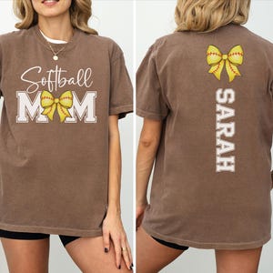 A woman wearing a brown t-shirt with the text "Softball Mom" and a yellow bow on the back.