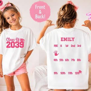 May include: White t-shirt with pink text that reads "Class Of 2039" and a graduation cap graphic. The back of the shirt has the name "EMILY" and grade levels from Pre-K to 12th grade. The shirt is worn by a young person.